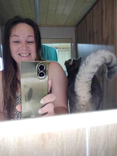 A woman is taking a selfie in a bathroom with a cat in the reflection of the mirror. The woman has long brown hair and is smiling. She is holding a smartphone in her right hand, which is capturing the selfie. The cat, which appears to be a husky or a similar breed with a distinctive fluffy tail, is standing behind her, partially visible in the mirror's reflection. The bathroom appears to be rustic, with wooden paneling on the ceiling and walls. The mirror is large and illuminated by a light strip at the bottom. The overall scene suggests a candid moment captured in a private setting. The location is identified as Vazery, Russia.