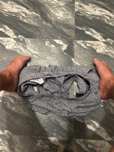 In this top-down shot, two feet, belonging to a person, are visible on a tiled floor. The floor has a marbled pattern with grey and white tones. In the center of the frame, a pair of grey and white striped boxer shorts are laid out, bunched up. The person's feet are positioned on either side of the shorts, with one foot holding down the waistband of the shorts. The lighting suggests an indoor setting, possibly a bathroom or bedroom, and it appears to be daytime. There are no other individuals or significant objects in the frame besides the feet and the shorts. The overall impression is a casual and perhaps intimate moment, as the shorts are presented with the feet. The location context is Kadewatta, Sri Lanka, but this information does not provide direct visual cues within the image itself.
