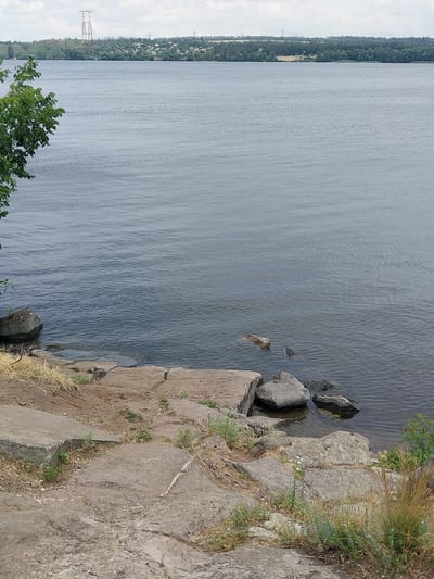 The image captures a serene riverfront scene in Liubymivka, Ukraine. The foreground is dominated by a rocky, uneven shoreline with several large stones and patches of dry grass and scattered green sprouts. Large, flat, grey concrete slabs are also present, suggesting a man-made modification of the natural landscape. The water of the river is calm, with a gentle ripple pattern across its surface. In the distance, across the river, a lush green treeline borders a settlement of small buildings, indicative of a village or town. A prominent, tall, lattice-like metal structure, possibly a transmission tower, stands on the horizon, silhouetted against a cloudy, overcast sky. The overall impression is one of a tranquil, somewhat rugged natural environment juxtaposed with signs of human presence.