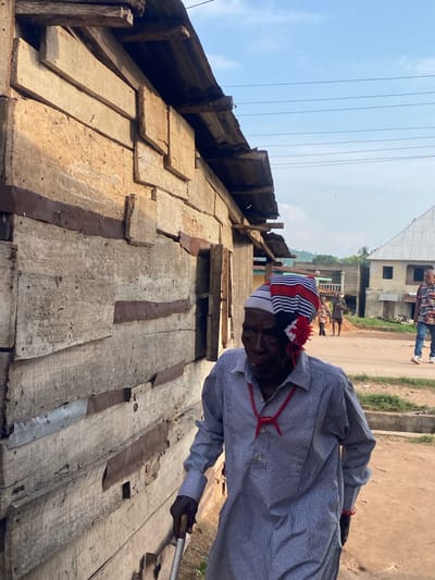 An elderly man, centrally positioned and walking from left to right, is the primary subject of this image taken in Igbara, Nigeria. He wears a long-sleeved blue and white checkered tunic, a red cord necklace with a knot, and a distinctive knitted cap featuring red, white, and blue stripes with a red floral-like embellishment on the side. His right hand holds a metal stick or cane, partially obscured.

To the man's immediate left, a building constructed from irregularly placed, weathered wooden planks secured by rusty metal bands forms a significant part of the foreground. The building's roof, composed of corrugated metal, is partially visible.

The scene unfolds on an unpaved, earthy street. In the middle ground, several other individuals are present; a man in a patterned shirt and blue jeans walks towards the right, and two more figures are visible further back. Various residential or commercial structures, appearing to be in different states of construction or disrepair, line the street. A distant landscape of rolling hills covered with sparse vegetation is visible under a light blue sky, which is traversed by multiple parallel overhead power lines.