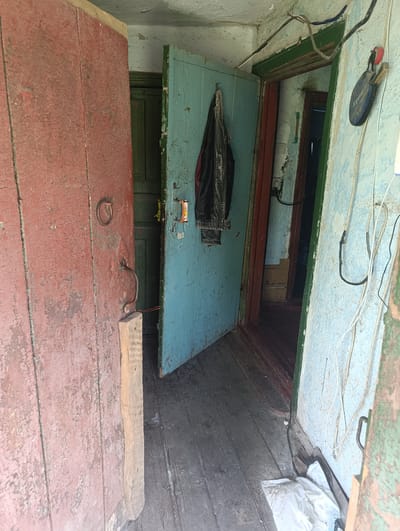 The image depicts an interior hallway with multiple doorways and worn wooden surfaces, likely in a residential setting. The scene is illuminated by natural light entering from an unseen source. The primary focus is on two adjacent doorways.
The left doorway, in the foreground, features a rustic, weathered red wooden door with peeling paint. A dark metal ring serves as a rudimentary handle, and a similar metal latch is visible. A short, light-colored wooden plank is propped against the bottom of this door.
The doorway to its right is slightly ajar, revealing a teal-colored door. This door also shows signs of wear and has a dark handle. A black jacket is hanging on this door. Behind the teal door, another doorway is visible, with a dark green door.
The hallway floor is made of aged, grey wooden planks. The walls appear to be plaster, with peeling paint in light blue and white hues. Electrical wires are strung along the walls and across the ceiling. A round, dark object with some labeling is attached to the wall near the teal door, possibly a sensor or a decorative item.
Further into the hallway, another doorway with a dark red frame is visible, leading to a darker interior space with a visible staircase. The overall impression is one of an older, possibly neglected, structure with a lived-in, utilitarian appearance. The image does not contain any specific markers or context that would allow for precise geolocation within Ladyzhyn, Ukraine, beyond the general impression of a residential interior.