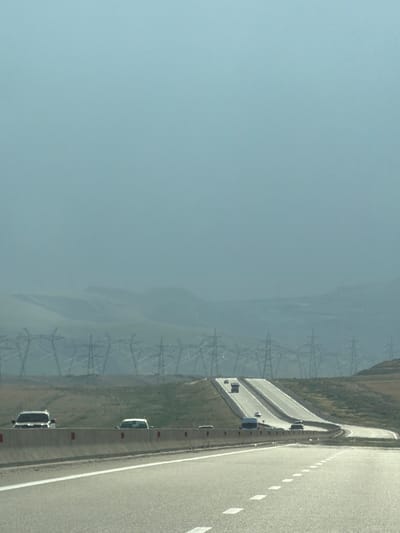 The image depicts a highway scene in the vicinity of Jangi, Azerbaijan. The highway itself is paved with distinct white dashed lines marking the lanes. A concrete barrier separates the lanes, with red reflectors visible on its surface. Several vehicles are present on the highway, including a white SUV, a white sedan, a dark-colored SUV, a dark-colored van, and other vehicles further in the distance. The landscape beyond the highway is characterized by rolling hills covered in dry grass. In the mid-ground, numerous tall power line towers stretch across the horizon, interconnected by wires, suggesting a significant power transmission infrastructure. In the background, distant mountains are shrouded in haze, with a pale, overcast sky overhead. The overall impression is that of a rural highway passing through a landscape with prominent industrial elements.