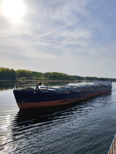 A medium shot depicts a large cargo barge, identified as "PAVEL LEBEDEV" in Cyrillic script on its bow, navigating a river. The vessel is long and flat-decked, laden with numerous rectangular cargo containers. It is painted in dark blue and red with a light-colored cabin structure towards the stern.
The surrounding environment features a calm body of water reflecting the sunlight. Lush, green trees line the banks of the river in the background. The sky is light blue with scattered clouds and a bright sun near the upper left corner of the frame. The photograph was taken in Ul'yanovo, Russia.