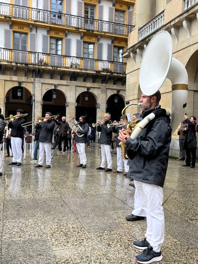The image shows a group of eight musicians in white pants and black jackets performing in the Plaza de la Constitución in Donostia-San Sebastián, Spain. The musicians are all wearing white pants, black jackets, and black shoes. They are playing a variety of instruments, including a trumpet, a tuba, a trombone, and a sousaphone. The musicians are standing in a line, facing the camera. The sousaphone player is in the foreground. He is wearing a black jacket over a white collared shirt and a black beanie, and he is playing a white sousaphone with a silver bell. His fellow musicians are dressed similarly. Behind the musicians are several people who are listening to them. There is a large building in the background. There are numbers on the building, numbered 144, 145, 146, 147, 98. The floor is made of stone and is wet, likely due to recent rain. There are several archways on the side of the building and chairs are present. The musicians are playing outside on a cobblestone square. It appears to be a public gathering or performance of some sort, with a crowd of people listening and observing.  The scene takes place in a square with a paved surface, which is partially wet. The architecture is classic, European, with a large, multi-story building in the background. The building has a yellow-brown color, and the windows have black shutters. The arched entryways with white painted edges open to the square.