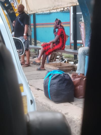 A man in a black polo shirt and shorts stands on a sidewalk, looking up. To his right, a woman in a red and black dress and headscarf sits on a bench. In front of her, a black bag with a blue strap and a brown bag rest on the ground. To the right of the woman, a person in light blue jeans sits on the bench. The background shows a colorful fence and some debris. The scene appears to be a bus stop in Oworonshoki, Nigeria.