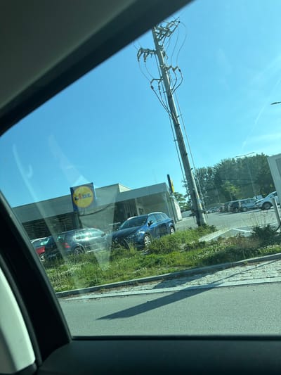 The image shows a view from a car looking out at a Lidl supermarket parking lot in Stanowice, Poland. The sky is clear and blue, with a utility pole with electrical wires prominently featured in the foreground. Several cars are parked in the lot, including a dark gray Volkswagen Golf and a blue Renault. The Lidl store itself is visible with its distinctive yellow and red logo. The parking lot is bordered by a grassy area with some wildflowers. The road in front of the parking lot is asphalt. The overall scene depicts a typical day at a suburban supermarket.