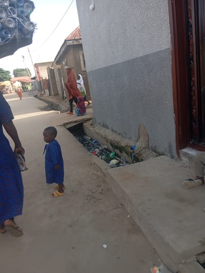 A medium shot shows a boy standing in a street with a building on the right. The boy is wearing a blue outfit and yellow sandals. A woman in a blue dress is partially visible on the left. The street is paved and has a drainage ditch filled with trash on the right side. In the background, other people and buildings are visible. The scene appears to be in a dusty, outdoor urban environment. The building on the right has a textured gray wall and a dark brown door frame. The overall impression is a snapshot of everyday life in a residential or market area.