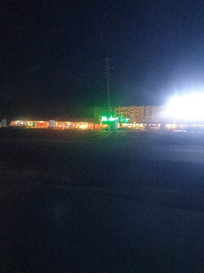 A dark image shows a line of lights dividing the frame vertically. The upper portion displays a bright, overexposed light source. Below this, a string of decorative lights descends, including a green cross or sign, leading to more indistinct orange and yellow lights. The background is largely obscured by darkness. The photo's orientation is skewed.