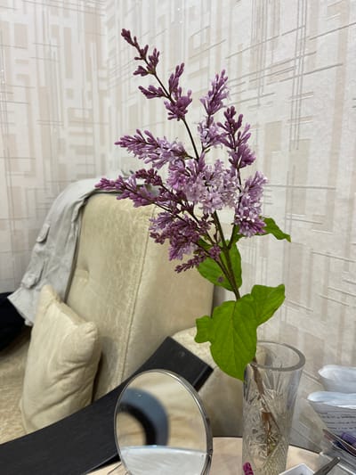 A sprig of light purple lilacs in a clear glass vase sits on a dark, reflective surface, likely a table. The vase is positioned to the right of center in the frame. In the background, a beige, upholstered chair with light-colored cushions is visible. A light-colored garment, possibly a jacket, is draped over the back of the chair. The wall behind the chair features a textured wallpaper with a geometric pattern of intersecting lines in a lighter shade of beige. To the left of the vase, a small, circular mirror with a silver rim is partially visible, reflecting a dark shape. To the right of the vase, stacks of papers and other small objects are present. The lighting appears to be artificial and somewhat diffused, casting soft shadows. There are no people or animals in the scene. The location is stated as Magdagachi, Russia.