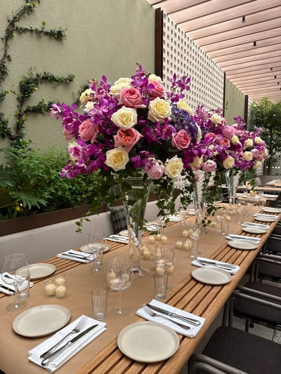 An outdoor or semi-outdoor dining area, likely a restaurant patio or event space, in West Palm Beach, United States of America, is prepared for a formal gathering. The central feature is a long, multi-segment wooden slatted table covered with a light brown paper runner. The table is meticulously set with numerous place settings, each consisting of a light beige plate (both round and oval), a folded white napkin, a fork, a knife, a clear glass wine goblet, and a clear glass tumbler. Small, lit votive candles in clear glass holders are distributed along the table's length.
Dominating the table are multiple large, elaborate floral centerpieces presented in tall, clear glass trumpet vases. These arrangements comprise a dense mixture of pink roses, cream/pale yellow roses, bright purple orchids, and blue-purple hydrangeas, with green foliage trailing from the vases.
The immediate environment includes a textured light olive-green wall on the left, partially covered by climbing green vines. Further back and to the right, a decorative white concrete block screen wall is visible, beyond which appears another section of the olive-green wall and lush green vegetation in planters. The ceiling overhead is constructed from light wooden slats, featuring several black recessed spotlights. Dark-framed chairs with black cushioned seats are arranged along the tables. No individuals are present in the scene.