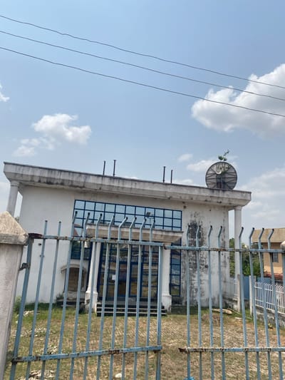 This image captures the exterior of a two-story building in Afikpo, Nigeria, observed from behind a metal fence. The primary structure is off-white with a flat roof featuring a pronounced, weathered cornice. The upper floor displays a series of large, rectangular windows with blue-tinted glass arranged in a grid pattern. Flanking the building are two classical-style columns, one partially visible on the left and another on the right. The exterior walls exhibit dirt, staining, and wear.
On the flat roof, several dark, cylindrical vents or pipes protrude vertically. To the far right of the roofline, a large, weathered, circular satellite dish is visible, with a small plant growing from its structure.
In the immediate foreground, a tall, light-blue painted metal fence with vertical bars and horizontal cross-sections stands, displaying significant rust and peeling paint. This fence partially obscures the lower section of the building and a patch of unkempt, dry grass and weeds between the fence and the building's facade.
Above the building, three parallel, dark utility wires traverse the upper portion of the frame. The sky is clear light blue with scattered white cumulus clouds. No individuals are present, and no discernible actions or interactions are occurring within the scene. To the far right, beyond the main building, another structure with a darker facade is partially visible.