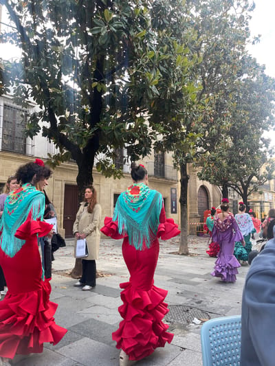 Several women are standing on a stone-paved sidewalk under a large tree in what appears to be Jerez De La Frontera, Spain. They are wearing traditional flamenco dresses in varying colors, including red and purple, with fringed shawls. Some have flowers in their hair. A woman in a tan trench coat is holding a white bag with black lettering. In the background, there is a stone building with arched doorways and windows. The scene captures a gathering or celebration in the city, showcasing its cultural traditions.