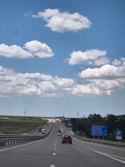The image shows a highway scene under a bright blue sky with scattered white clouds. The highway has multiple lanes and is lined with a metal guardrail. Several vehicles are visible on the road, including cars, trucks, and a van. The landscape adjacent to the highway consists of grassy hills and trees. There are also signs indicating directions to Simferopol, Yevpatoriya, and Sevastopol, along with distances. A parking sign is also visible. The photo appears to be taken from a moving vehicle, looking forward along the highway. The location is identified as Трудовское сельское поселение, Ukraine.