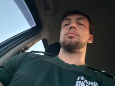 A close-up, low-angle, point-of-view shot captures a young adult male sitting inside a vehicle. The man has short dark hair, a light brown beard, and his eyes are closed. He is wearing a dark green t-shirt with white lettering visible on the chest area. The interior of the vehicle is dimly lit, with a portion of a sunroof visible at the top left of the frame, allowing natural light to enter. The scene appears to be in the city of Varvara, Bulgaria.