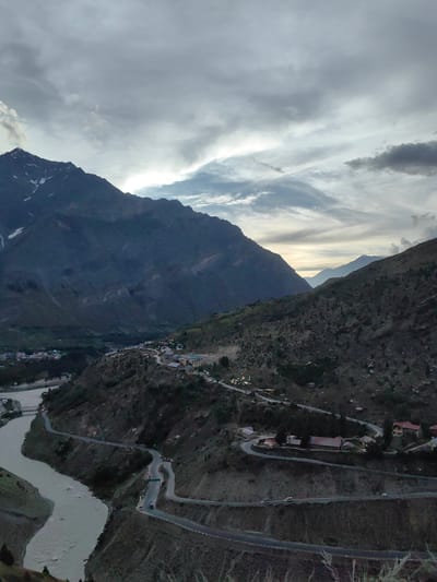 This is a high-angle, wide shot of a mountainous landscape in Gushal, India, likely taken during dusk or dawn. The sky is overcast with dramatic, wispy clouds, suggesting impending rain or recent storm activity. Dominating the scene are large, dark mountain ranges, some with snow-capped peaks visible, casting shadows over the valley below.

A wide, muddy river snakes through the valley floor, its banks lined with sparse vegetation. A winding road with multiple hairpin turns carves its way up the side of the mountain. Along this road, small clusters of buildings, possibly a village or a collection of guesthouses, are nestled into the hillside. A few vehicles are visible on the road, suggesting human activity and transit. The overall mood is serene and awe-inspiring, with the rugged natural beauty of the mountains juxtaposed with the man-made elements of the road and settlement. The lighting is subdued, characteristic of twilight, and the shadows add depth and drama to the rugged terrain. There is no visible text in the image.