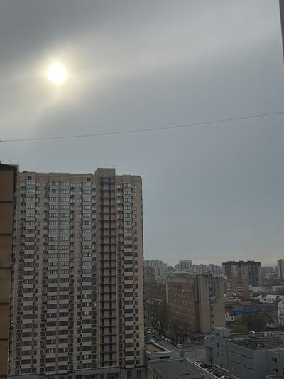 The image depicts a cityscape in Kuleshovka, Russia, under a hazy sky with the sun visible through the overcast. A large, tan-colored building with numerous windows dominates the foreground. Adjacent to it and continuing into the distance are various other buildings, forming a dense urban landscape. A thin wire stretches across the upper portion of the frame. No people or specific actions are visible.