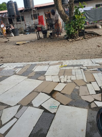 The image depicts an outdoor scene within a residential area in Isunba, Nigeria. The immediate foreground features an irregularly laid pavement comprising various shapes and sizes of light grey and beige concrete slabs interspersed with darker gravel and dirt. A discarded yellow strip and a piece of green and white litter are visible on this surface, with a partial view of a black tire in the bottom right corner.

In the mid-ground, the ground transitions to packed dirt. To the far left, a woman in a multi-colored patterned top and skirt stands near a small, light-colored table with items on its surface. Centrally, a second woman, wearing a light-colored top, a patterned wrapper, and a dark headwrap, operates a red engine-powered grinding machine, likely for foodstuffs, mounted on a black metal table. Several black plastic buckets are positioned around and beneath this machine. To the right of the grinding operation stands a prominent tree trunk with some green foliage emanating from its base and lower sections.

In the background, behind the women, are single-story residential structures with corrugated metal roofs. A number '3' is visible above a doorway on one building. Two large black plastic water tanks are elevated on a metal scaffold structure behind the buildings. Other elements in the background include a black plastic bin, clothes hanging on lines, and a red cloth item. The overall environment appears to be an open-air compound or yard.