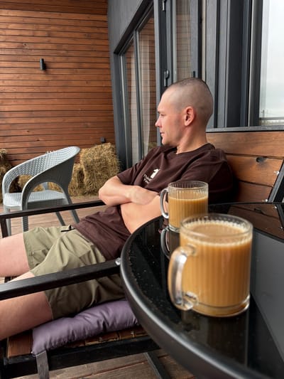 A man with a shaved head sits with his arms crossed on a wooden bench outside a building with a wooden facade and large windows. He is wearing a brown sweatshirt and khaki shorts. In front of him, on a black glass-top table, are two clear glass mugs filled with a light brown liquid, likely coffee or tea. A grey wicker chair and hay bales are visible in the background. The scene appears to be an outdoor seating area.