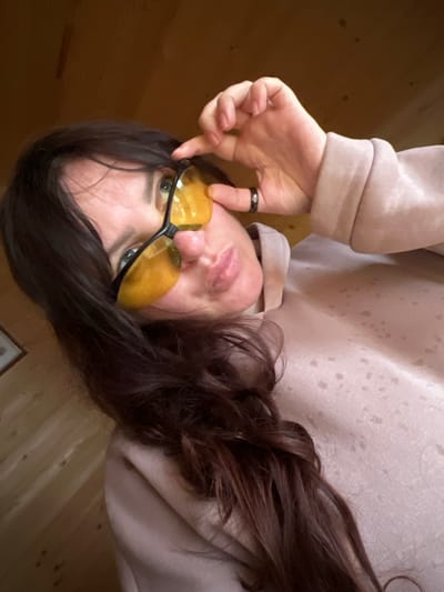 An eye-level, close-up shot captures an individual, presumably an adult female, looking directly at the viewer with her head slightly tilted. Her long, dark brown hair cascades around her. She is wearing a light beige or pale pink hooded sweatshirt. A pair of yellow-tinted glasses, possibly for night driving or blue light filtering, covers her eyes, with her right index finger touching the frame as if adjusting them. A black ring with subtle lighter speckles is visible on her right index finger. Her lips are slightly parted, suggesting a subtle expression. The background features warm-toned wooden paneling, indicating an indoor setting, possibly a room or cabin in Protasy, Russia. The lighting is soft and diffused, characteristic of indoor daytime conditions. There is no visible text in the image. The overall impression is casual and contemplative.