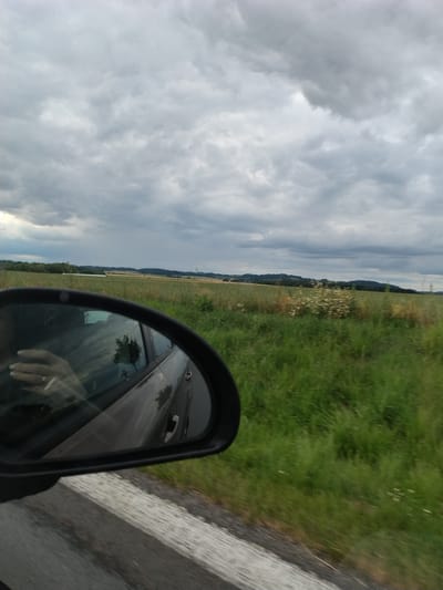 The image shows a view from a car's side mirror, looking out at a rural landscape under a cloudy sky. The mirror reflects the interior of the car, including a person's hand holding a cigarette. The landscape features fields of green grass and some scattered white flowers, with a line of trees and rolling hills in the distance. The location is specified as Drhovice, Czechia. The road visible below the mirror has a white stripe and a textured concrete barrier. The overall impression is one of a journey through a countryside on an overcast day.