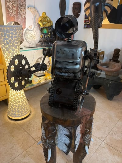 This indoor shot depicts a cluttered display in what appears to be a shop or market setting, potentially in Grau I Platja, Spain, considering the query. The scene is dominated by a steampunk-style lamp made of dark metal with gears, chains, and cylindrical components, resting on a patterned, illuminated base. The lamp is surrounded by various curios and decorative items, including a wooden sculpture of elephants arranged in a row along a dark wooden plank. Other objects include decorative wooden carvings, what appear to be African masks, and a golden Buddha statue. The items are placed on surfaces with neutral colors, likely tiled or wooden, adding to the overall eclectic and somewhat cluttered environment. The setting suggests a place where unique, handcrafted items are sold.