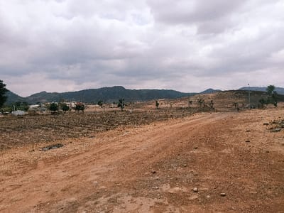This image presents an expansive outdoor scene in Rafin Pa, Nigeria, under an overcast sky filled with grey clouds. The foreground is dominated by a wide, reddish-brown unpaved dirt road, which tracks from the lower center towards the mid-distance on the right. The ground immediately adjacent to the road consists of parched, reddish-brown earth with sparse, dry vegetation and scattered small stones.
In the mid-ground to the left, a cluster of light-colored, multi-story buildings is visible among green trees. Adjacent to these structures are white tanks or containers, and a utility pole stands upright. Further across the mid-ground, the landscape is uneven and arid, characterized by dry soil, scattered small trees with minimal foliage, and multiple dark utility poles extending into the distance. The background features a continuous range of dark, undulating hills or mountains beneath the cloudy horizon. No individuals or specific activities are discernible within the frame.