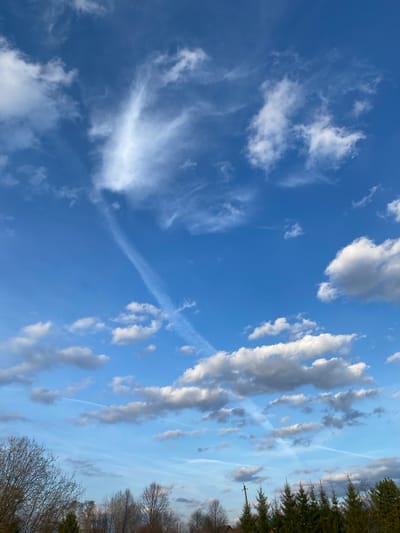 The image presents a daytime view of the sky, likely taken in or near the area of Ореховая Гора, Russia, based on the prompt. The sky is primarily a clear, vibrant blue, with scattered cumulus and cirrus clouds. Some of the clouds appear to be elongated, possibly due to wind shear. There are also noticeable contrails, indicating the recent passage of aircraft. The lower right corner shows the edges of trees, some with bare branches, suggesting it could be early spring or late autumn. The photo is taken from an upward-tilted angle, showcasing the vastness of the sky.