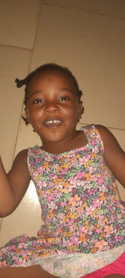 A young girl with brown skin and braided hair is lying on a light brown tiled floor. She is smiling and wearing a floral print dress with pink shorts underneath. The scene appears to be indoors. The exact location in Ashipa, Nigeria, is not discernible from the image.