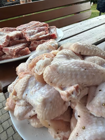 A close-up shot showcases two plates of raw meat, likely prepared for grilling, placed on a wooden picnic table in the city of Mordarka, Poland. The plate in the foreground is filled with raw chicken wings, seasoned with pepper. Behind it, a second plate holds what appears to be raw pork ribs, also seasoned. The background shows a wooden bench and glimpses of a garden setting with greenery. The lighting suggests it's daytime, possibly late afternoon or early evening. No people are clearly visible, though a blurred figure might be present in the upper left corner. The ground beneath the table is paved with grey cobblestones.
