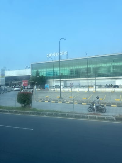 The image shows a view of a large, modern building with a prominent "cinépolis" sign, indicating it is likely a cinema complex. The building features a green-tinted glass facade and a white upper section. In front of the building, there is a parking lot with cars and a motorcycle. A person is riding a motorcycle in the foreground, moving from left to right across the image. A road is visible in the bottom portion of the frame, with a white line marking the lane. The overall environment appears to be urban, with a clear sky overhead. The presence of the Cinépolis sign and its typical architectural style suggest a location within a city. Based on available information, Cinépolis has a presence in various cities in Indonesia, including Serang. Therefore, this scene is likely located in Serang, Indonesia, and depicts the exterior of a Cinépolis cinema and its surrounding area.