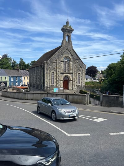 In Donegal, Ireland, a stone church with a bell tower stands prominently against a blue sky with wispy clouds. A gray sedan drives past the church, reflecting its facade on its hood. Behind the church, colorful houses line a street, and trees provide a lush green backdrop. A black truck is parked near the houses. The scene is set on a sunny day, with a clear view of the architecture and surrounding urban landscape.