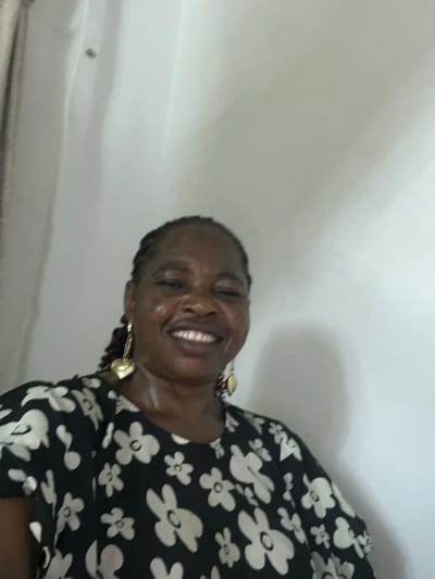 A dark-skinned woman is smiling at the camera. She is wearing a black and white floral dress and gold earrings. The background is a white wall. This image was likely taken indoors. It is not possible to determine the location in the city of Mopo-Akinlade, Nigeria, from the information provided.