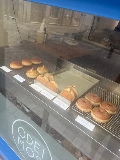 A display case of donuts is visible through a glass window. The donuts are arranged on metal racks within the display. Some donuts are plain, while others are topped with meringue, cream, or covered in a glaze. White labels with descriptions and prices are placed in front of some of the donuts. In the background, reflections of a city street and buildings are visible. A blue and white logo with the text "ODE MO" is printed on the glass in the lower left corner of the image. This logo, "Odei Mo," is associated with a donut shop located in Granada, Spain.