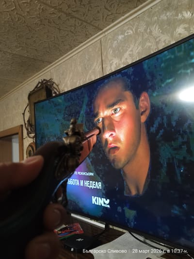 The image depicts an indoor scene in Balgarsko Slivovo, Bulgaria, captured on March 28, 2026, at 10:37 AM. The foreground features a light-skinned human hand holding an antique-style, dark-gripped pistol with brass-colored ornamentation. The pistol's barrel is directed towards a large, curved television screen positioned in the midground. The television displays a close-up of a man's face with short dark hair and light eyes, illuminated by a source from the right side of the screen. Cyrillic text, including "ТЕ РЕЖИСЬОРИ," partial "БОТА И НЕДЕЛЯ," and "KINO NOVA," is visible on the screen, which exhibits digital pixelation. In the background, a room interior is visible with a textured ceiling and patterned wallpaper on the walls. A decorative object, resembling an abstract wall clock or sculpture with dark, twig-like elements, is mounted on the wall to the left of the television. A wooden door frame is partially visible on the far left. Below the television, a dark-colored "A1" branded set-top box or media player rests on a surface, beside which a white piece of paper is visible.
