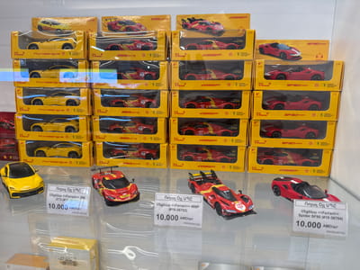 The image shows a retail display of toy cars, specifically model Ferraris. The cars are arranged in clear plastic packaging on yellow cardboard boxes, stacked on shelves. The majority of the boxes have "Shell V-Power" branding, indicating they are likely promotional items. The specific models visible are Ferrari Purosangue, 296 GT3, 499P, and SF90 Spider, all in 1/41 scale and rated for ages 6+.
In the foreground, three individual toy cars are displayed outside their packaging, each with a price tag. The price tags are written in Armenian script and show a price of 10,000 AMD/шт (Armenian Dram per unit). The cars in the foreground are a yellow Ferrari Purosangue, a red and yellow Ferrari 296 GT3, and a red Ferrari SF90 Spider. The overall scene suggests a toy store or a promotional display within a larger retail environment. The lighting is bright, suggesting an indoor setting during daylight hours. There are no people visible in the image. The background shows more shelves with similar toy car packaging, extending the display further back.