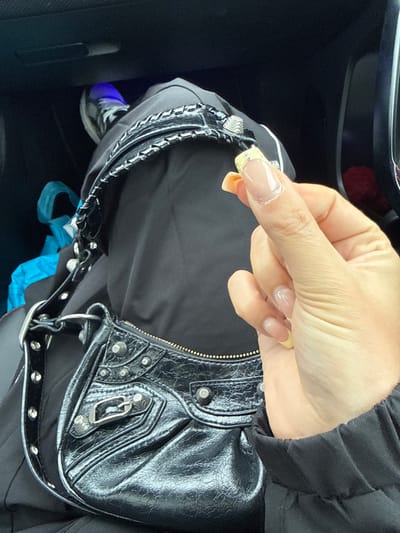 The image depicts the interior of a vehicle. A human subject's right hand is visible in the foreground, holding a small, light-colored, indistinct object between the thumb and forefinger. The fingernails on the visible hand are manicured in a French style, featuring translucent pink/nude bases and light yellow tips, with a small, circular decorative element affixed to the tip of the index finger. The subject's right arm is partially visible, clad in a black long-sleeved garment. A black, textured handbag, featuring silver-toned metal studs, buckles, and zippers, and a braided strap, is positioned in the central-lower frame, resting on what appears to be the subject's lap or an adjacent seat. In the background, to the left, a portion of the subject's leg, dressed in black fabric, is visible, alongside a dark athletic shoe with a white sole. A crumpled blue plastic bag is visible on the floorboard in the lower-left quadrant. A blue ambient light emanates from a lower-left area of the vehicle's interior. The specific location within Sint-Niklaas, Belgium, cannot be determined from the provided visual information.