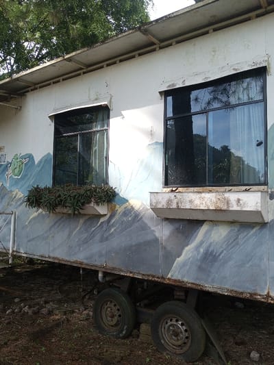 A white mobile home rests on dual axles with rusty wheels. The exterior features two black-framed windows, one with a planter box overflowing with greenery. The walls are painted with a mountain scene in shades of blue, grey, and white. The flat roof has visible supports. The home is situated outdoors, surrounded by sparse vegetation and dark soil.