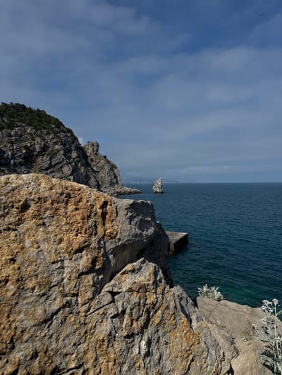 A view from a cliffside in the Гаспринский поселковый совет, Ukraine shows a rocky coastline meeting the sea. The foreground is dominated by rough, textured rocks in shades of gray and brown, some with sparse white plants clinging to their surfaces. The sea stretches into the distance, appearing calm with a dark blue hue. A small rock formation rises from the water closer to the shore. The sky is partially cloudy with patches of blue.