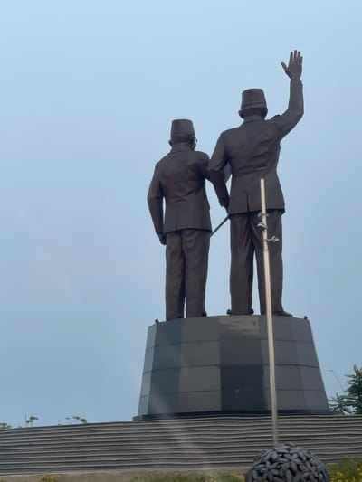 The image depicts a monument of two men in suits and hats, seen from behind, against a hazy, pale blue sky. They are standing on a dark, octagonal pedestal. The man on the right has his right arm raised in a waving gesture. A metallic pole with several clips is positioned in front of the two figures. Below the pedestal are wide, tiered steps made of dark material, leading down towards a foreground with some low-lying plants and a dark, spherical sculpture. The environment appears to be outdoors, possibly in a public space, with a general sense of overcast weather. The location is specified as the city of Kosambi Barat, Indonesia.
