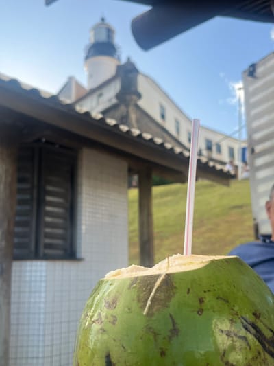 The image depicts a green coconut, partially cut at the top with a white and red striped straw inserted, in the foreground. The coconut is slightly out of focus. In the midground, a low building with a tiled roof and white square tiled walls is visible on the left, featuring a louvered window. Behind this structure, the distinctive white Farol da Barra (Barra Lighthouse) with its dark top stands prominently against a bright blue sky. Adjacent to the lighthouse complex, a multi-story building with white walls and numerous windows is situated on an elevated, grassy embankment. A partial view of a person wearing a dark blue shirt is visible on the far right. The scene is outdoors on a clear day, located near the Farol da Barra in the city of Salvador, Bahia, Brazil.