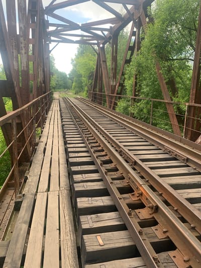 The media file shows a perspective view along a weathered railway bridge, oriented vertically in the frame. The bridge is constructed of rusted metal girders and wooden planks, with parallel sets of train tracks running along its length. The tracks themselves are also aged, showing signs of rust and wear. The scene is framed by lush green trees and foliage on either side, with a bright, overcast sky visible in the distance. The perspective draws the eye down the length of the bridge, creating a sense of depth and leading towards a vanishing point where the tracks disappear into the greenery. There are no people visible in the media file. The overall impression is one of an abandoned or disused railway line, possibly in a rural or natural setting. The weather appears to be overcast or hazy, as indicated by the bright but diffused light. The location cues suggest a place with ample vegetation and a significant railway infrastructure, consistent with the context of Kokhma, Russia. The image does not contain any visible text.
