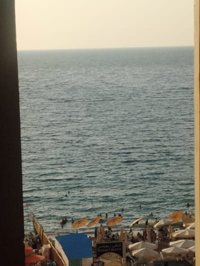 The image captures a view from a building overlooking the Mediterranean Sea, specifically the beach area known as Regency Beach in Alexandria, Egypt (latitude: 31.273, longitude: 30.001). The sky is clear, the sea is a calm blue, and the sand is visible along the shore. There are people swimming in the sea, sunbathers lounging on beach chairs, and numerous yellow umbrellas scattered across the beach. The image is framed by a window, with a wooden beam to the right and a dark wall on the left. A blue roof and a black sign reading "Regency Beach" in Arabic and English are visible in the foreground. The scene is peaceful and relaxed.