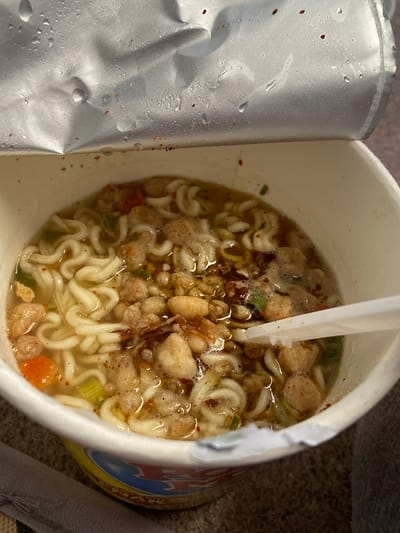 A close-up, overhead shot shows an opened cup of instant noodles, with the foil lid peeled back and condensation droplets visible. The noodles are submerged in a broth, and pieces of dried vegetables, possibly carrots and scallions, are scattered throughout. Some small, brownish pieces, possibly meat or crispy toppings, are also present. A white plastic fork is partially submerged in the noodles. The cup itself is white with some colorful graphics on the lower portion, although the text is not legible. The background appears to be a textured surface, possibly a table or floor. The lighting suggests an indoor setting. There is no discernible indication of the location being Sekar Mawar, Indonesia.