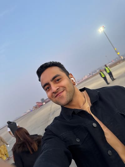 A man takes a selfie outdoors with an expansive backdrop. He wears a dark jacket and has short, dark hair. An earbud is visible in his ear. In the background, people in yellow safety vests stand near a light pole. A clear, light sky stretches above them. The photograph is rotated counter-clockwise, creating a tilted perspective. Other people can be seen in the background.