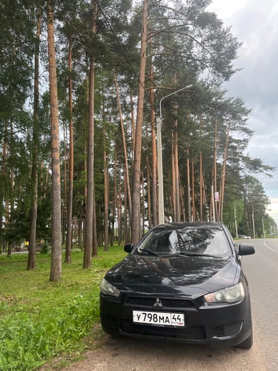 An eye-level, medium shot captures a black Mitsubishi Lancer parked on the side of a road, bordered by a dense forest of tall pine trees. The car is positioned at an angle, facing slightly left, with its front bumper and license plate clearly visible. The license plate reads "Y 798 MA 44" and has "РОССИЯ" written below it, indicating its Russian origin. The road to the right of the car is asphalt and curves away into the distance. In the background, to the left of the trees, a children's playground with yellow structures and red picnic tables can be seen. The sky is overcast with grey clouds. The overall scene is in the city of Nikolskoye, Russia.