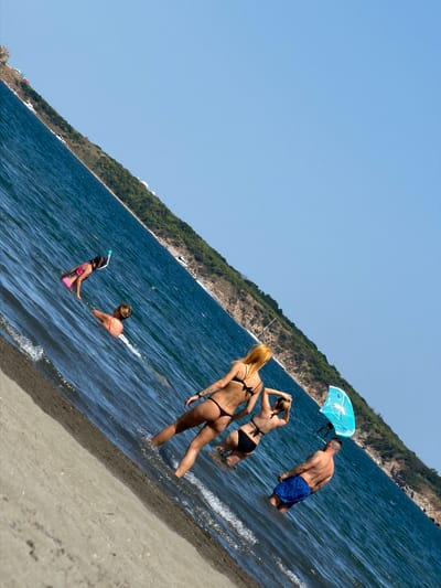 The image depicts a group of people on a beach in an unknown location in Bulgaria. The scene is shot with a diagonal perspective, making the waterline appear to tilt. In the foreground, a sandy beach meets the calm, blue water.
Several individuals are visible in the water. To the left, a young person wearing a pink swimsuit and snorkel gear is partially submerged. Further back, a person in an orange swimsuit is swimming. In the middle of the frame, two women in bikinis are wading in the water. One woman with blonde hair is facing away from the camera, while the other is facing towards the water. To the right, a man wearing blue patterned swim shorts stands in the water. A bright blue and white surfboard is positioned near him. In the background, a green, tree-covered hillside slopes down towards the water, with some white buildings or structures visible along its edge. The sky is clear and blue.