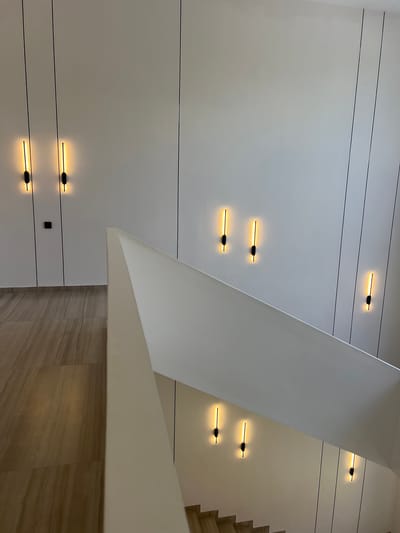 This is an interior shot of a modern staircase in Pitsunda, Abkhazia. The walls are white with thin black vertical lines accentuating the architecture. Several minimalist sconces with vertical yellow lights are mounted on the walls, illuminating the space. The staircase itself is made of wood with a white railing. The flooring on the lower level is tiled with a wood-like pattern. There are no people visible in the image.