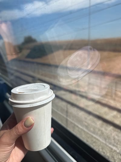 A medium shot captures a person's hand holding a white paper coffee cup with a white plastic lid. The cup appears to be near a window with a partial reflection of the cup itself visible on the glass. Outside the window, the background shows a blurred view of what appears to be building exteriors with a pattern of light and shadow, possibly due to sunlight filtering through window blinds or structures. The photograph was taken in the city of Modi'in-Maccabim-Re'ut, Israel.