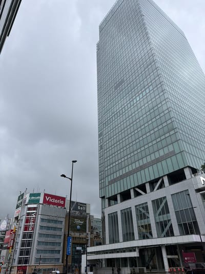 This is a medium-shot, eye-level, exterior photograph of the cityscape in Shinjuku, Japan. The sky is overcast and gray. The primary subject is a very tall, modern skyscraper with a glass facade, positioned on the right side of the frame, taking up a significant portion of the right half of the image. The skyscraper's glass panels are a pale blue-green hue, and the building has a grid-like pattern. Below the main glass section of the skyscraper, there is a lower section with large windows and geometric structural elements.

To the left of the skyscraper, a cluster of mid-rise buildings with prominent signage is visible. These buildings feature a mix of architectural styles and present various advertisements, including a large red and white sign that reads "Victoria," another green and white sign for "L-Breath," and a sign for "Victoria Golf." Some of these buildings lean slightly, giving them a dynamic appearance. In the foreground, there are streetlights, traffic signals, and pedestrian areas. A few cars are parked on the street, and a few individuals are visible walking or standing on the sidewalk. The overall impression is an urban environment with a mix of contemporary and older commercial architecture under a cloudy sky.