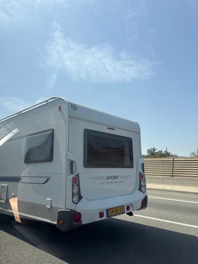 The image captures the rear view of a white caravan with the model designation "SPORT NL-PREMIUM LINE" visible. It is on a paved road, likely a highway, given the presence of a concrete barrier with horizontal slats and a chain-link fence behind it. The sky is clear blue with sparse white clouds. The caravan has a license plate displaying "54-WD-DX" and what appears to be a "100" sticker, indicating a speed limit. There is a reflection of the sky and possibly trees on the caravan's windows. The environment suggests it is a sunny day in a rural or semi-rural setting. The scene is static, with no people or dynamic actions depicted. The location is identified as Maureillas-las-Illas, France.